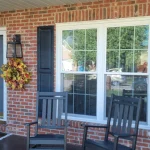 A large white-framed window on the front of a red-brick home. The window shutters and front door are black, as is the set of rocking chairs on the porch right in front of the window.