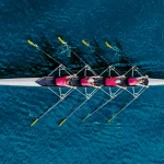 Aerial view of four rowers in matching pink shirts paddling in unison across deep blue water in a racing shell.